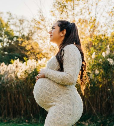 Pregnant woman in a white dress poses outdoors, surrounded by greenery and soft sunlight.