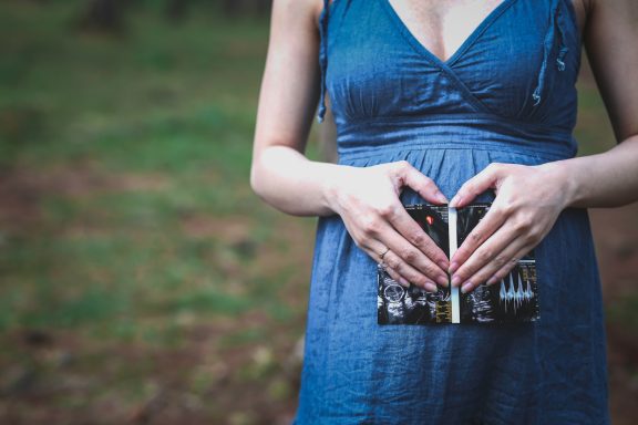 A person in a blue dress holding a book close to their belly, forming a heart shape with hands.