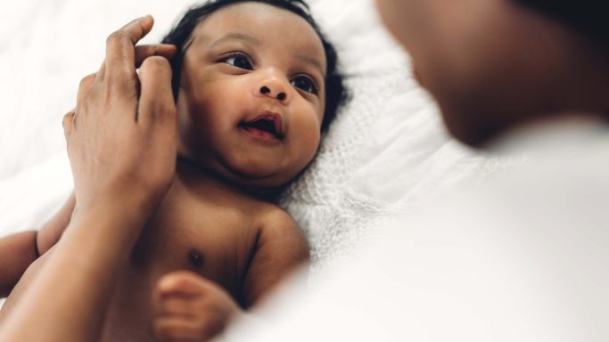 A close-up of an infant looking curiously at an adult's hand gently touching their face.