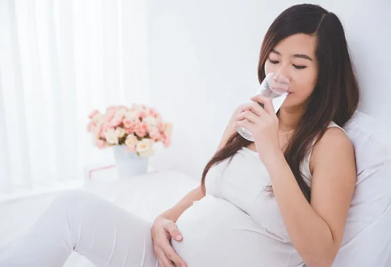 Pregnant woman sitting on a bed, drinking water, with flowers in the background.